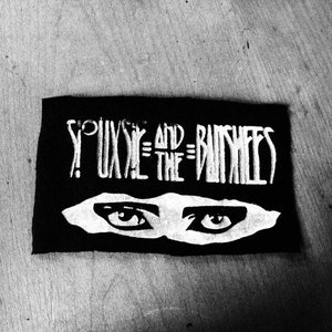 May include: Black and white patch with the text "Siouxsie and the Banshees" and a pair of eyes.