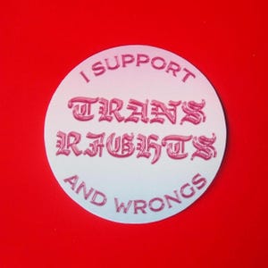 Puede incluir: Una pegatina circular blanca con un degradado rosa y el texto "I support trans rights and wrongs" en una fuente estilizada.
