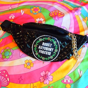 Puede incluir: Riñonera negra acolchada con una cadena dorada y un parche que dice "BODILY AUTONOMY FOREVER" en verde y blanco. La bolsa está sobre una colorida tela de patrón psicodélico con setas y flores.