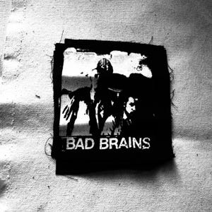 Peut inclure: Patch thermocollant noir et blanc avec un logo de groupe. Le patch représente une silhouette d'une personne avec le texte "BAD BRAINS" en dessous.