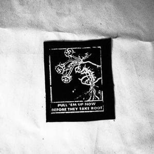 以下が含まれることがあります： 「PULL 'EM UP NOW BEFORE THEY TAKE ROOT」というテキストと、植物を持った手のグラフィックが描かれた、白黒の刺繍パッチです。