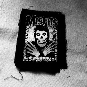 Pode incluir: Patch bordado preto e branco com a palavra "Misfits" em letras brancas e uma figura com rosto de caveira com uma mão esquelética branca.