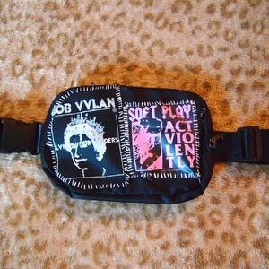 Könnte beinhalten: Schwarze Bauchtasche mit schwarzem Gurt. Die Tasche hat zwei aufgenähte Patches. Ein Patch hat ein Schwarzweißbild einer Person mit dem Text "Bob Vylan" und der andere Patch hat rosa Text, der "Soft Play Act Violently" lautet.