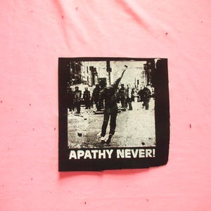 Puede incluir: Parche en blanco y negro que representa una escena callejera con una persona lanzando un objeto. El texto "APATHY NEVER!" está impreso en blanco en la parte inferior. El parche es cuadrado y está sobre un fondo rosa.