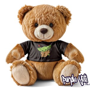 Op de afbeelding: Een bruine teddybeer met een zwart t-shirt met een groene cartoonfiguur. De beer heeft een zacht, pluche uiterlijk en zit rechtop. De afbeelding bevat de tekst "Purple Yeti Trading Co."