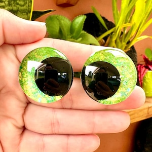 Könnte beinhalten: Zwei grüne und schwarze Plastikaugen mit Glitzerakzenten. Die Augen sind rund und haben eine schwarze Pupille.