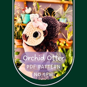 May include: A crocheted plush otter with a dark brown body, a cream-coloured face, and a light pink flower accessory on its head. The otter has large, dark, glossy eyes. Text on the image reads: Orchid Otter, PDF PATTERN, NO SEW.