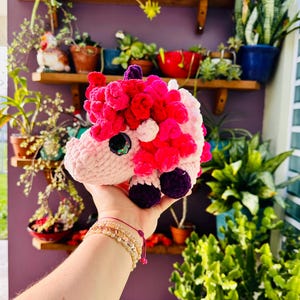 May include: A plush unicorn toy with a pink body, a mane of red and pink yarn, and purple hooves. The unicorn has a sparkly black eye and is held up in front of a background of plants and shelves.