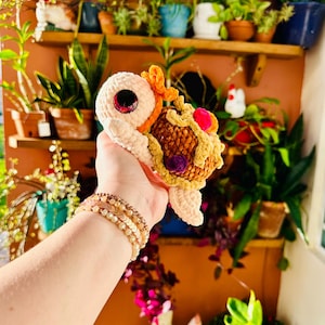 Puede incluir: Una tortuga de ganchillo hecha a mano con cuerpo beige, caparazón marrón y ojos rojos grandes. La tortuga tiene detalles en naranja y rosa en el caparazón y la cabeza. Se sostiene frente a un fondo de plantas y estanterías.