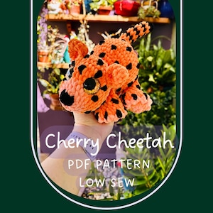 May include: A plush crochet cheetah toy made of soft orange yarn with black spots, large dark eyes, and a small black nose. The text on the image reads Cherry Cheetah PDF PATTERN LOW SEW, presented in a white font against a purple overlay.