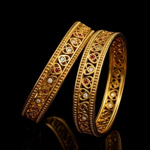 May include: Two gold bangles featuring a repeating openwork heart pattern along the center. Each heart is accented with either a small round clear stone or a small round red stone. The edges of the bangles are finished with a beaded texture.