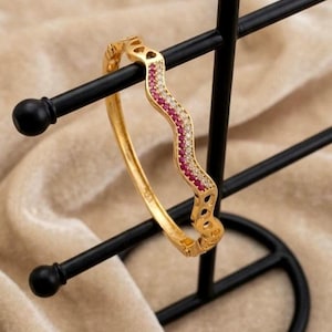 Könnte beinhalten: Ein goldfarbener Klapparmreif mit wellenförmigem, geschlängeltem Design. Die Oberfläche ist mit einer zentralen Reihe kleiner, leuchtend rosa Steine verziert, die von zwei Reihen klarer, funkelnder Steine in einem polierten Metallrahmen flankiert werden.