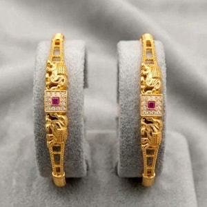 May include: A pair of gold bangles featuring an intricate elephant motif on each side of a central square setting. The square center contains a single square-cut red gemstone surrounded by small clear stones, with openwork metal detailing.