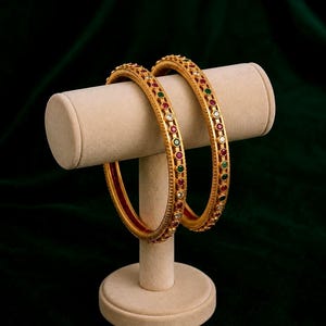 May include: Two gold bangles with detailed patterns and set with red, green, and white gemstones, presented on a cream stand. The bangles have a polished finish and a traditional design, suitable for adding a touch of elegance.