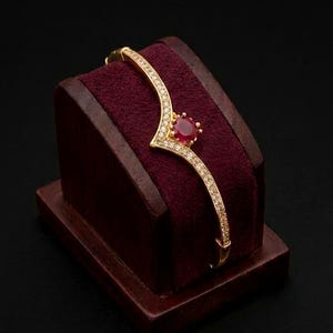 May include: A gold-colored bracelet with a V-shaped design, adorned with small, clear gemstones. The center features a large, oval-shaped, red gemstone. The bracelet is displayed on a burgundy velvet jewelry stand.