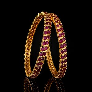 May include: Two gold bangles, each adorned with a repeating pattern of oval, ruby-coloured gemstones. The bangles are set against a black background, which accentuates the jewellery's design and colour. The bangles are likely made of gold.