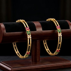 May include: Two gold bangles with colourful gemstone accents, displayed on a dark wooden stand. Each bangle features a repeating pattern of red, green, and white stones. The stand has a dark velvet surface.