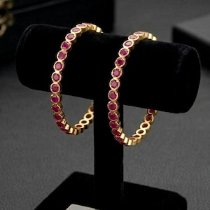 May include: Two gold-colored bangles with a continuous row of red gemstones are displayed on a black velvet stand. The bangles are circular and have a repeating pattern of the red gemstones. The stand is on a black surface.