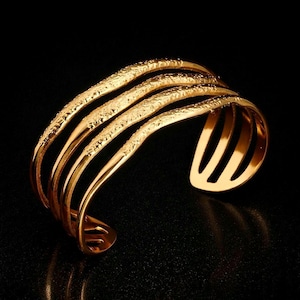 May include: Gold-coloured cuff bracelet with a textured surface. The bracelet features five parallel bands, each with a detailed pattern, curving to create an open design. The jewellery is set against a black background.