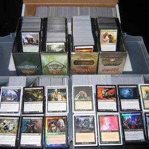 May include: A collection of Magic: The Gathering trading cards, organized in a clear plastic storage container. The cards are in various colors and feature fantasy artwork. Several card boxes are visible, each labeled with the game's logo.