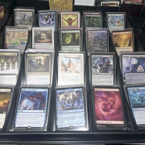 May include: A black plastic storage tray containing multiple Magic: The Gathering trading cards organized in rows. Each card is protected by a clear plastic sleeve, displaying various fantasy illustrations, text, and game symbols on their surfaces.