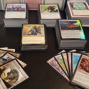 May include: A collection of Magic: The Gathering cards, including a stack of cards with a dragon illustration, a stack of cards with an angel illustration, and a stack of cards with a creature illustration. There are also several individual cards spread out on a black surface.