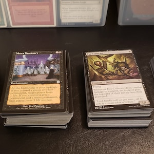 May include: Four stacks of Magic: The Gathering trading cards. Each card features unique artwork and text. The cards are black with various illustrations and text in white and other colors. The cards are stacked on a dark surface.