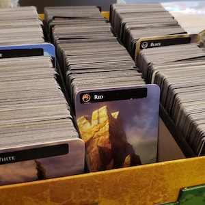 May include: A box filled with stacks of trading cards, organized by color. The colors are white, blue, black, red, and green. Each stack has a card on top with the color and a symbol printed on it.