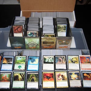 May include: A gray plastic storage box with multiple decks of Magic: The Gathering cards. The cards are organized in clear plastic sleeves and are stored in boxes labeled "Magic: The Gathering".