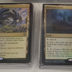 May include: Two Magic: The Gathering cards, Thran Portal and Voldaren Estate. Thran Portal is a gold and silver land card with a circular gate design. Voldaren Estate is a gold and black land card with a dark castle design. Both cards have the Magic: The Gathering symbol in the bottom right corner.