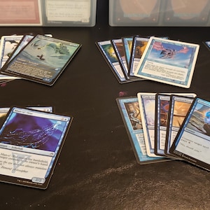 May include: A collection of Magic: The Gathering cards, featuring various blue and white cards with illustrations of creatures, spells, and lands. The cards are arranged in stacks on a dark surface.