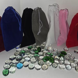 May include: Eight velvet drawstring pouches in burgundy, blue, black, gray, white, pink, red, and purple arranged behind a scattered collection of small, round, translucent glass gems in shades of clear, green, and blue.