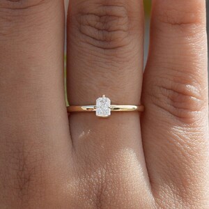 Puede incluir: Un delicado anillo de oro con una gema rectangular y transparente. El anillo tiene una banda fina y pulida, y la gema está engastada en un diseño simple y elegante. El anillo se muestra en una mano con esmalte de uñas de tono neutro.