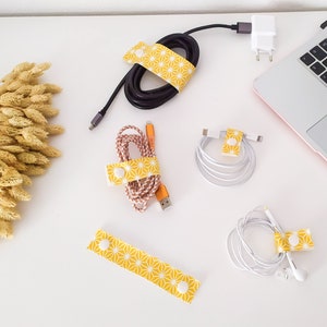 May include: Five yellow cable organizers with a white starburst pattern. The organizers are wrapped around different cords, including a black charging cable, a white charging cable, and a white headphone cord.