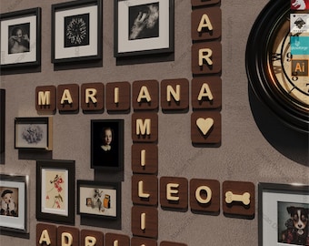 May include: A wall decor set featuring wooden Scrabble tiles spelling out the names "Mariana", "Leo", and "Adrian". The tiles are arranged in a crossword pattern with a heart-shaped tile in the center. The names are surrounded by framed pictures and a clock.