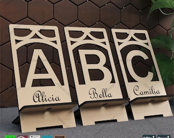 Puede incluir: Tres soportes de teléfono de madera con las letras A, B y C recortadas. Cada soporte tiene un nombre debajo de la letra: Alicia, Bella y Camilia. Los soportes son de color beige con bases negras y recortes decorativos en la parte superior. El texto "PHONE STAND TEMPLATE" está en la parte superior.