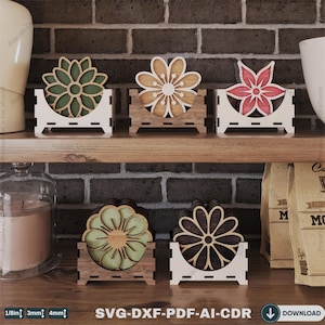 May include: Four sets of coasters with floral designs in green, brown, red, and black. Each set is displayed on a white wooden stand with a brown wooden base. The stands are arranged on a wooden shelf with a brick wall background.