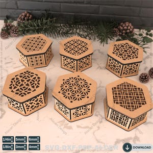 Peut inclure: Cinq boîtes hexagonales en bois avec des motifs découpés au laser complexes. Les boîtes sont en bois brun clair et ont une bordure noire autour des bords. Les boîtes sont disposées en demi-cercle sur une surface en bois.