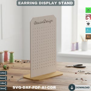 Puede incluir: Un expositor de pendientes de madera con múltiples agujeros para colgar pendientes. El expositor tiene forma rectangular con una base dorada. El texto "EARRING DISPLAY STAND" está en la parte superior, y "AtacanDesign" está impreso. El grosor es de aproximadamente 6mm.