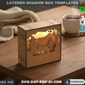 May include: A wooden layered shadow box with a forest scene illuminated from within. The box features a cutout design of trees and a wolf. The text "LAYERED SHADOW BOX TEMPLATES" and "SVG-DXF-PDF-AI-CDR DOWNLOAD" are visible.