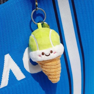 Puede incluir: Llavero de peluche con forma de cono de helado de tenis. La parte superior es verde claro con un diseño de pelota de tenis, con un borde blanco y esponjoso y una cara sonriente. El cono es de color canela y texturizado. Unido a un llavero plateado.