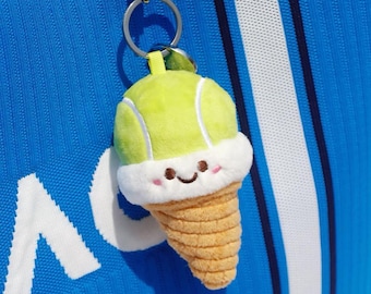 May include: A novelty keychain featuring a tennis ball ice cream cone design. The top is a light green tennis ball shape, with a white fluffy trim and a smiling face. The cone is tan and textured. Attached to a silver keyring.