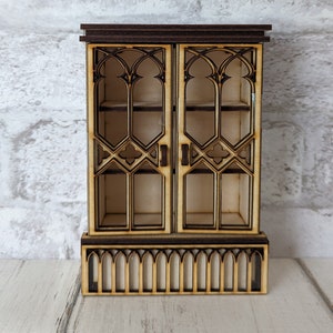 Op de afbeelding: Een klein houten miniatuurkabinet met een donkerbruine bovenkant en een decoratief, gotisch ontwerp. Het kabinet heeft twee glazen deuren met ingewikkeld traliewerk en een basis met bogen.