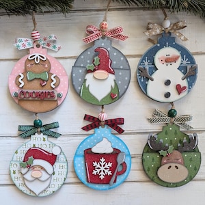 May include: Six wooden Christmas ornaments with festive designs. The ornaments feature a gingerbread man, gnome, snowman, Santa, hot cocoa, and a moose. Each ornament is round with a decorative ribbon and bead for hanging. The word "COOKIES" is on one.