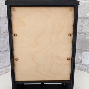 Puede incluir: Vitrina de madera negra con un panel trasero de madera clara y un estante. La vitrina tiene una parte superior decorativa y est&aacute; hecha de madera.
