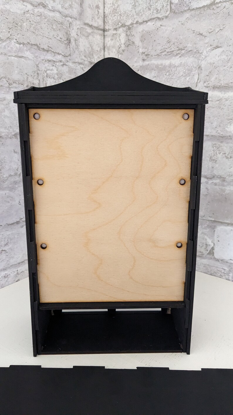 Puede incluir: Vitrina de madera negra con un panel trasero de madera clara y un estante. La vitrina tiene una parte superior decorativa y est&aacute; hecha de madera.