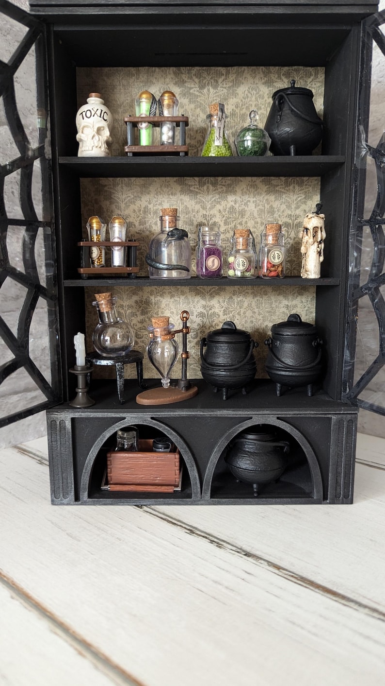Puede incluir: Un armario de madera negro con puertas de vidrio y estantes llenos de frascos de pociones, calderos y otros art&iacute;culos de bruja. El armario tiene una botella con forma de calavera etiquetada como "Toxic".