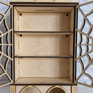 Puede incluir: Armario de madera con un pentagrama y un dise&ntilde;o de luna creciente en la parte superior. El armario tiene tres estantes y una secci&oacute;n inferior con dos aberturas arqueadas. El armario est&aacute; hecho de madera clara y tiene una mancha marr&oacute;n oscura.