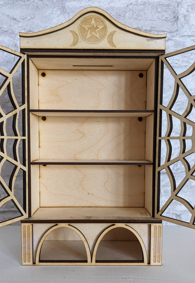 Puede incluir: Armario de madera con un pentagrama y un dise&ntilde;o de luna creciente en la parte superior. El armario tiene tres estantes y una secci&oacute;n inferior con dos aberturas arqueadas. El armario est&aacute; hecho de madera clara y tiene una mancha marr&oacute;n oscura.