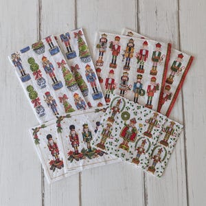 May include: A collection of white paper napkins with festive Christmas designs. The napkins feature illustrations of nutcrackers, Christmas trees, and decorative topiaries in various colors. Some napkins have red borders.